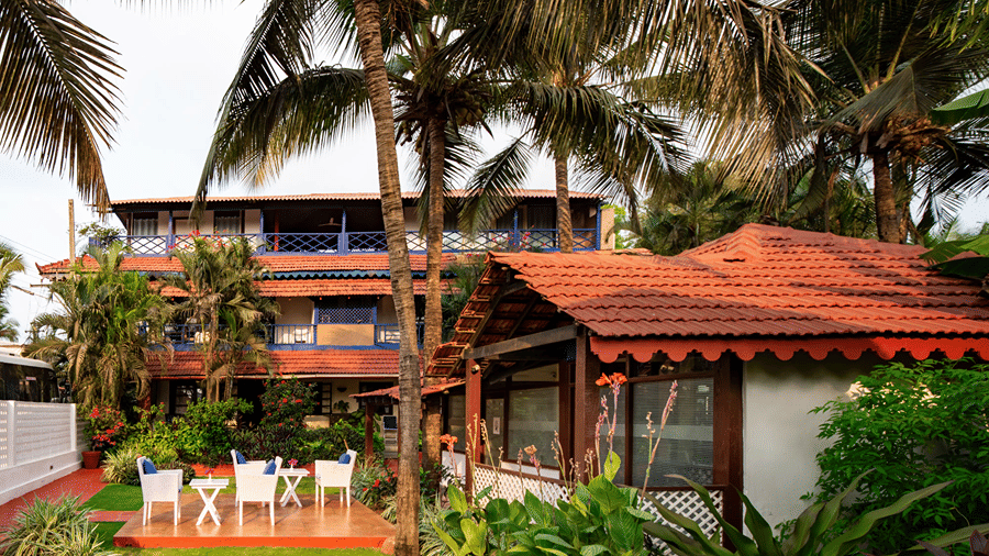 Red-roofed outdoor cottage surrounded by palm trees, tropical plants and garden seating.
