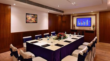 Board room with royal blue seating arrangement, bright lighting, flat screen TV, and wooden flooring.