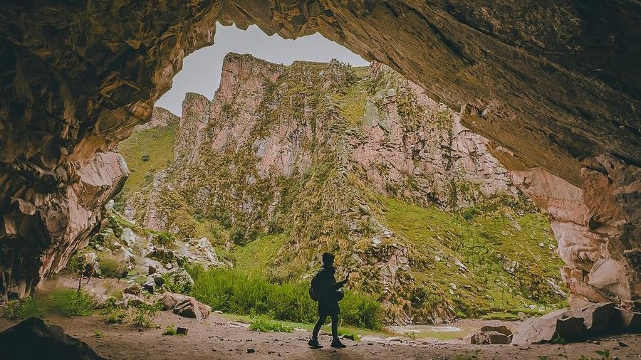 An image of a man in standing on the entrance of the cave which is surrounded by a rocky hill