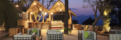 Outdoor seating area at dusk with cushioned chairs and sofas, illuminated by hanging lights under thatched-roof structures, surrounded by trees and a scenic mountain view in the background - Suryavilas Luxury Resort and Spa