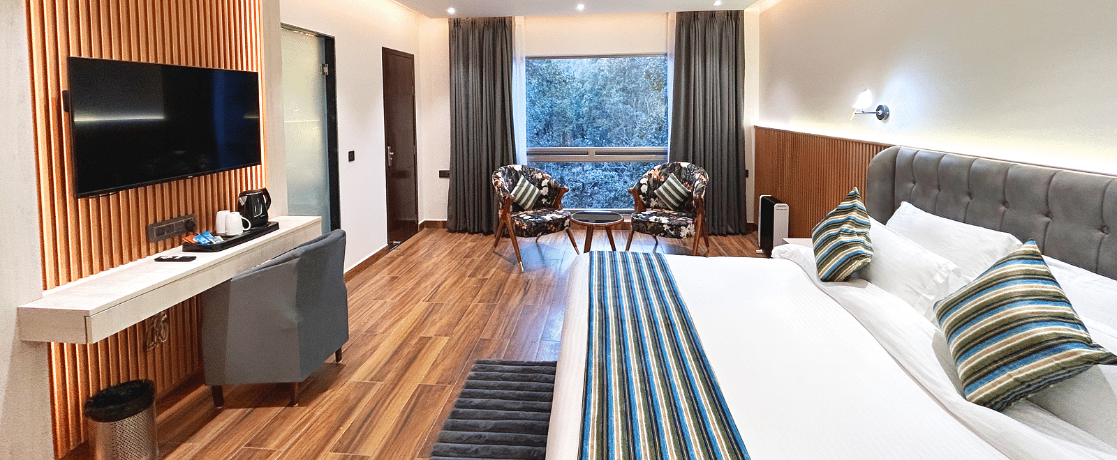 An image of the premium room at DLS The Emerald, Nainital featuring a bed, a flat screen TV, chairs and table and a window overlooking the view