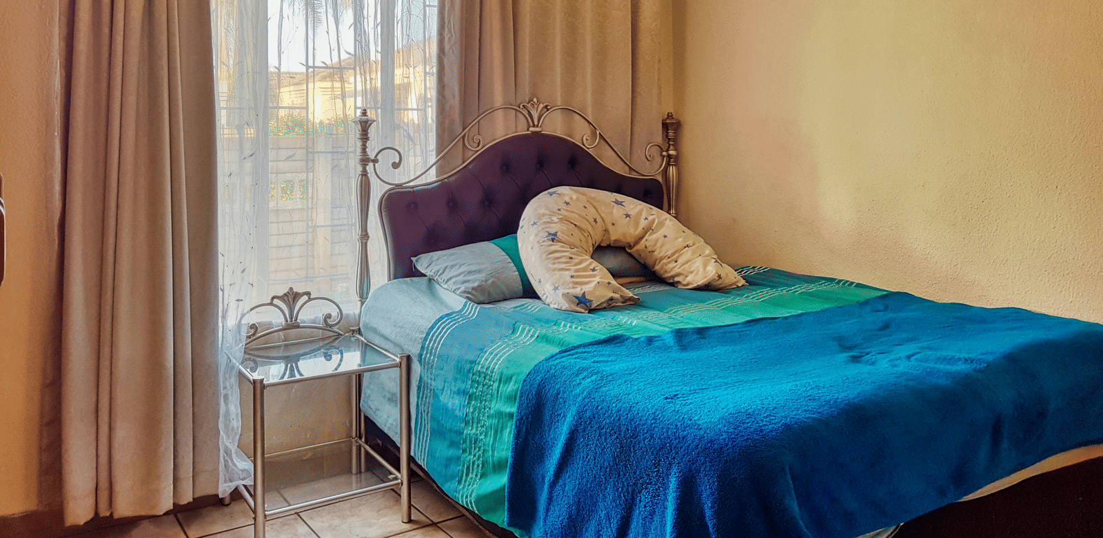 A bedroom featuring a double bed with blue sheets spread neatly over it with beige curtains covering the window behind it.