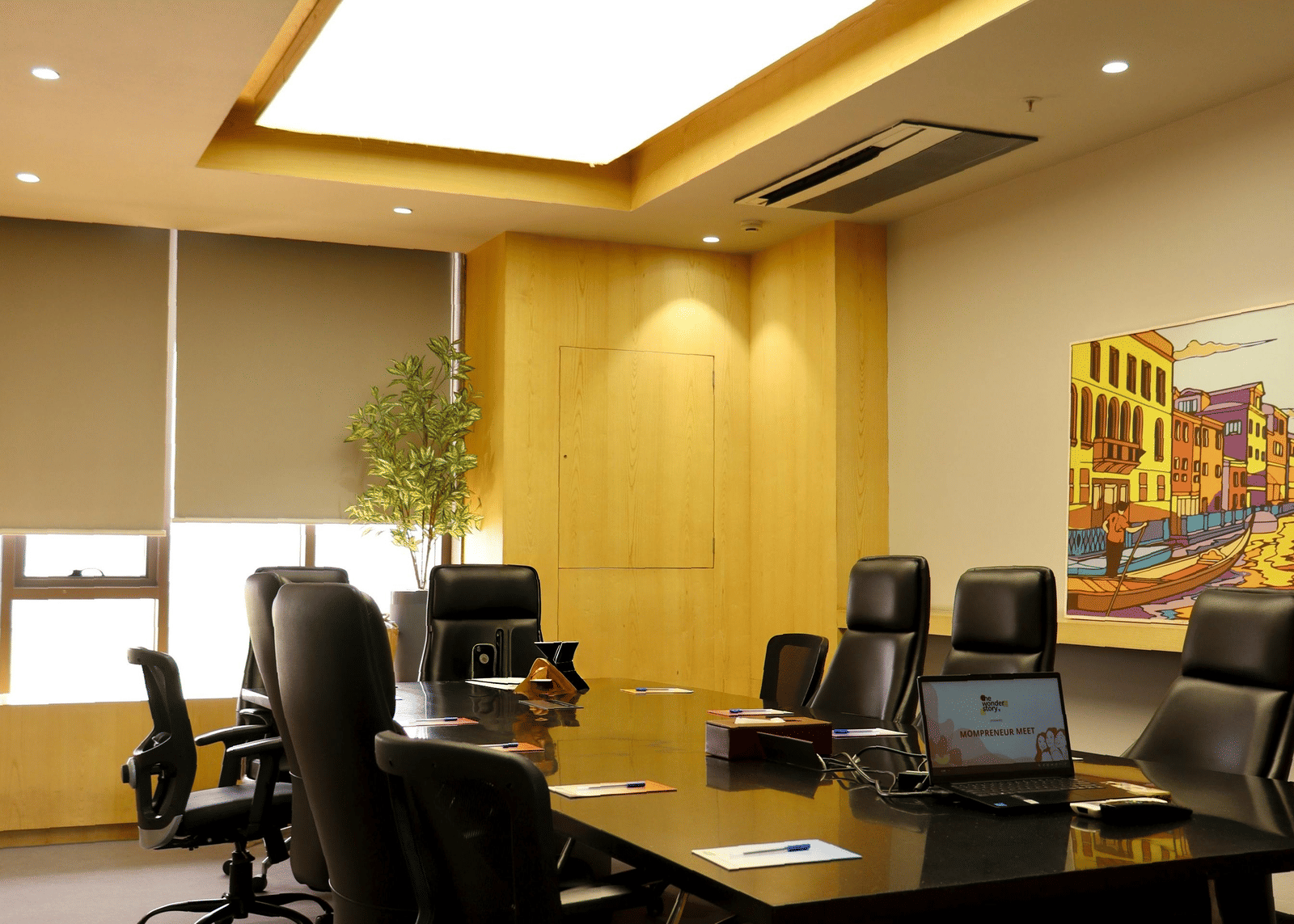 A modern conference room with a large wooden table, black office chairs, warm lighting, and a minimalist decor setup.
