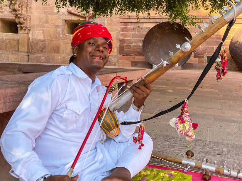 A man wearing a red turban and white clothing sits on a colourful mat outdoors, playing a traditional stringed musical instrument.