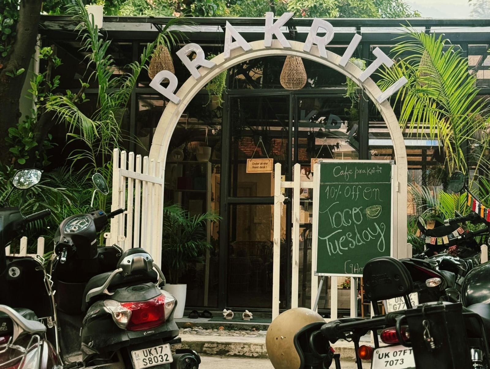 An entrance of a cosy cafe surrounded with green trees and bushes, with several two-wheeler vehicles parked outside.