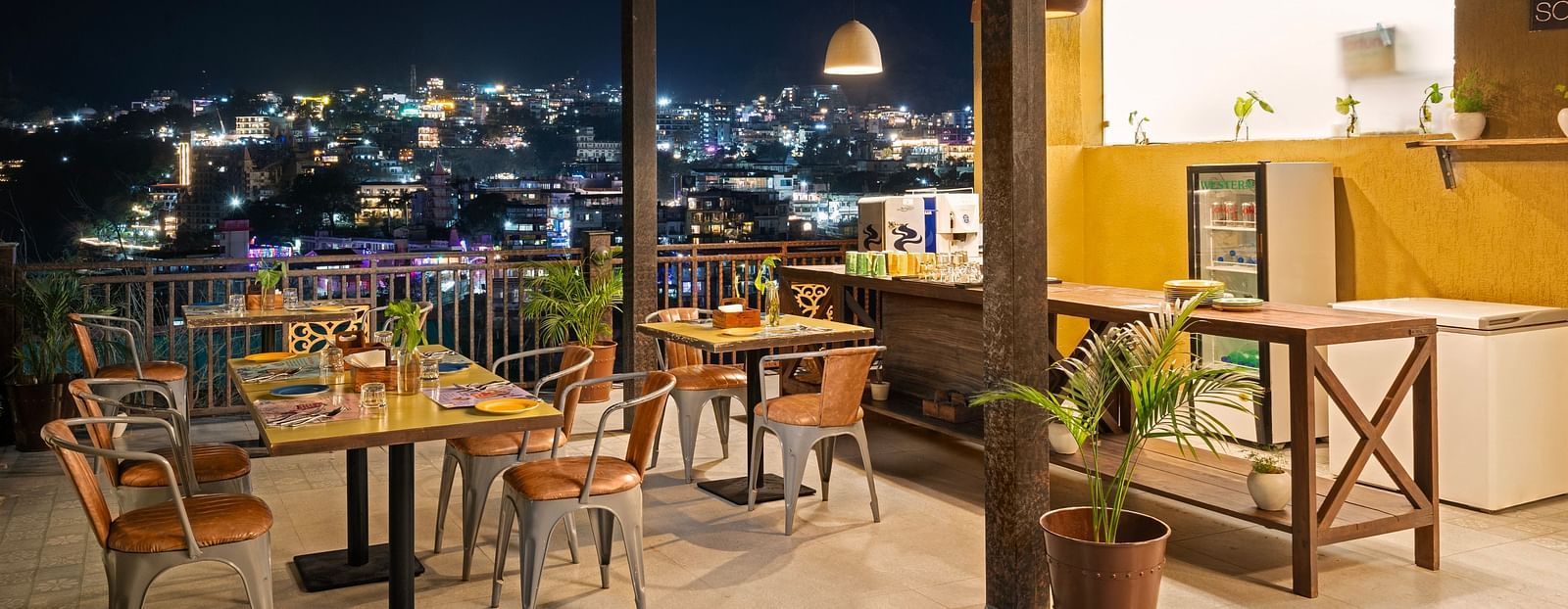Panoramic view of city skyline from rooftop restaurant, with tables, chairs, and city lights at night at Bedzzz, Rishikesh 