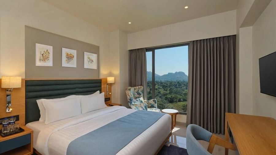 Side view of a room with a double bed and tree frames hanging on a wall and a balcony with scenic view outside an armchair and a tabe at Pride Resort Ambaji