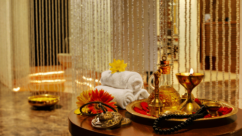 Myra Spa at Kenilworth Resort & Spa, Goa, featuring rolled white towels, a brass tray with massage oil, a lit lamp, fresh flowers on a round wooden table, and shimmering beaded curtains in the background.