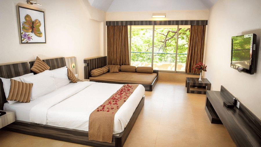 The Regency Deluxe room of the Adamo The Resort featuring a king size bed, a diwan on the side, a wall mounted TV, a centre table, a console, and floor to ceiling windows that lead to the balcony. 