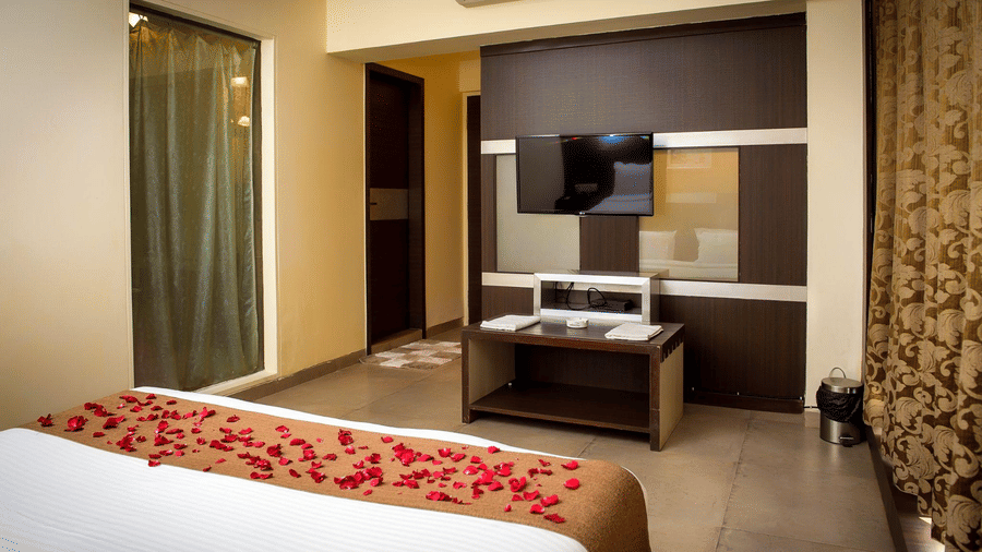 A hotel room with brown and yellow decor, a neatly made bed decorated with flower petals, a wall mounted TV, and a console.