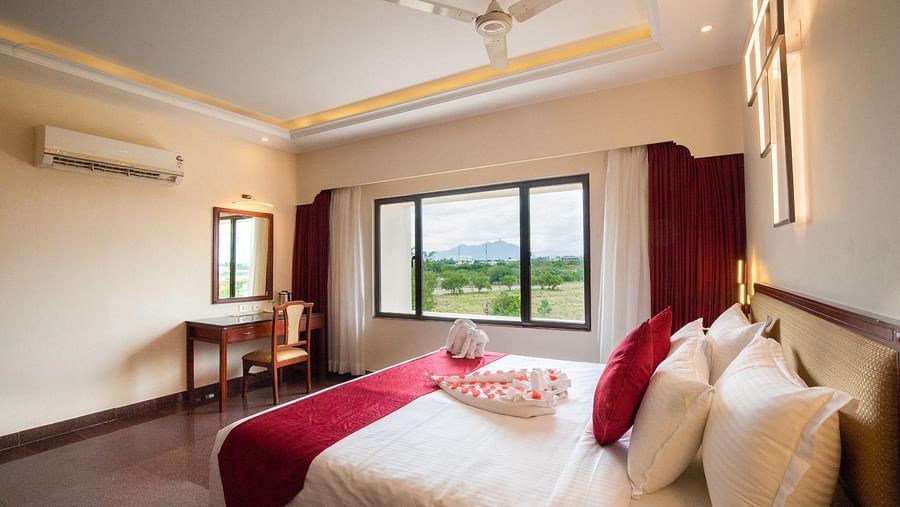 A hotel bedroom with a large bed, white linens, red accents, and an air conditioning unit.