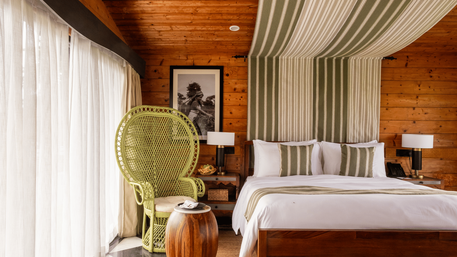 A rustic yet modern bedroom with a large bed, featuring a striped green and white canopy over the headboard. A bright green wicker peacock chair sits next to a large window with sheer curtains. The room has wooden paneled walls and ceiling, and a checkered