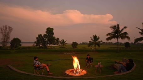 Campfire with chairs arranged around it on a grassy field, with a few people sitting and palm trees in the background.