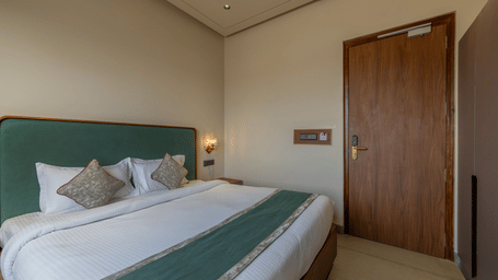 View of a room at Hotel Highway Prince, Behror, showing a large bed with a teal headboard and runner, a wooden door, and a wall-mounted light switch panel.