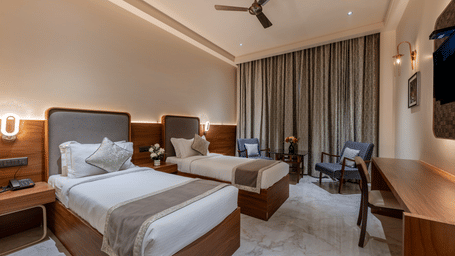 A twin bedroom featuring two beds with white linens, a ceiling fan, and dark curtains in the background.
