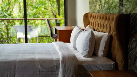 A close-up of a neatly made bed with white linens and a brown tufted headboard next to a window at Stanley Revelation.