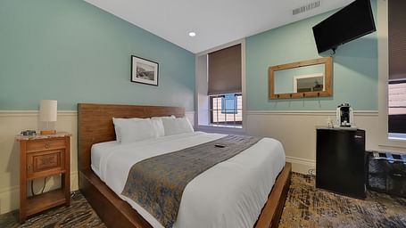 A comfortable room at The Groveland Hotel with sage green walls, wooden bed, blue patterned runner, TV, and convenient coffee station.