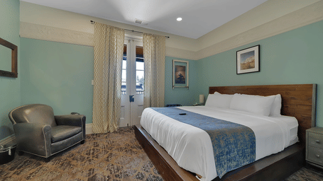 The Groveland Hotel's elegant room includes sage green walls, wooden platform bed, leather armchair, and French doors opening to a balcony.