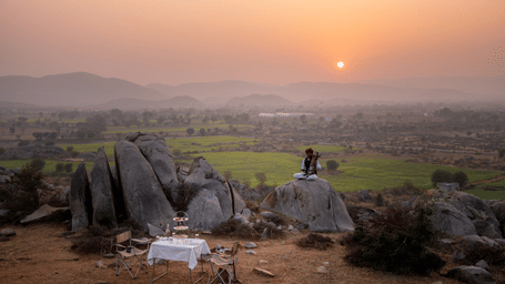 A folk musician sits on a boulder playing an instrument near a sunset dining setup overlooking fields at Utsav Camp Sariska.