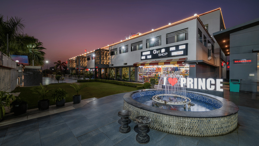 Hotel Highway Prince Paota circular fountain with I Love Prince branding at dusk, surrounded by lit retail outlets and twilight purple sky.