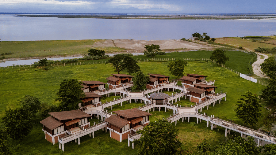 An aerial perspective of the interconnected wooden villas at Rhino & River Wildlife Retreat & Spa, Pobitora, joined by elevated walkways on a green lawn next to a river.