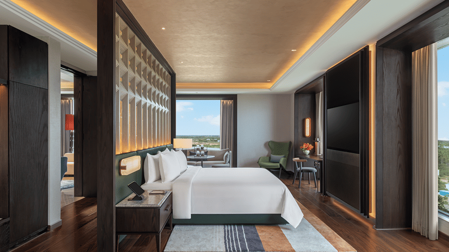 Presidential Suite main bedroom with a double bed against a carved partition, a wall-mounted TV, a desk and a green armchair at The Residency Signature Madurai.