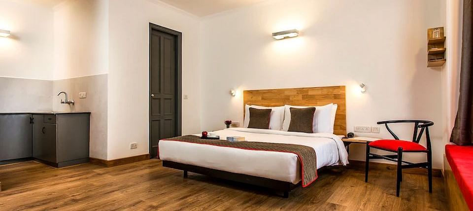 A spacious hotel room with a wooden bed, side chair and kitchenette with neutral tones - TGI Ameya Grand, Goa