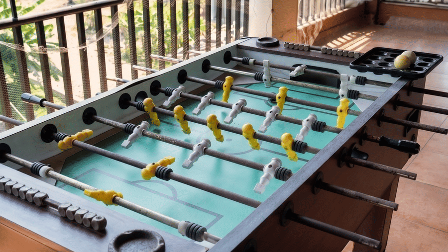 A foosball table rests on a tiled terrace balcony, featuring metal railings, green foliage in the background at Bougainvillea, Alibaug.