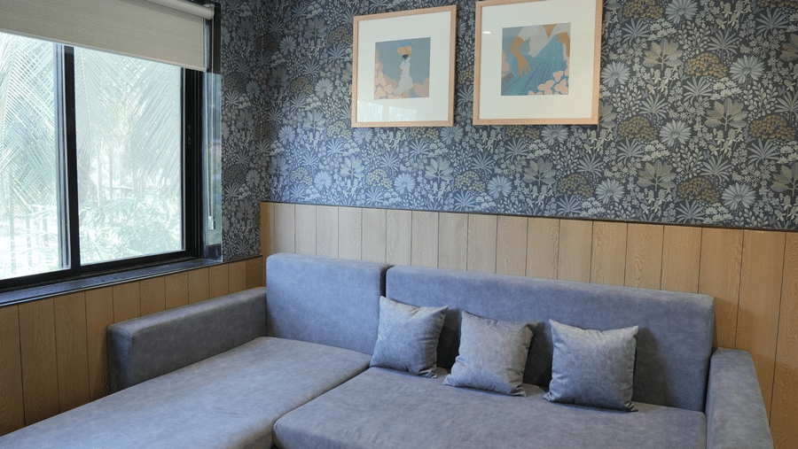 An L shaped sofa bed sits against a patterned wallpapered wall adorned with 2 framed pictures under a white ceiling fan at Bougainvillea, Alibaug.
