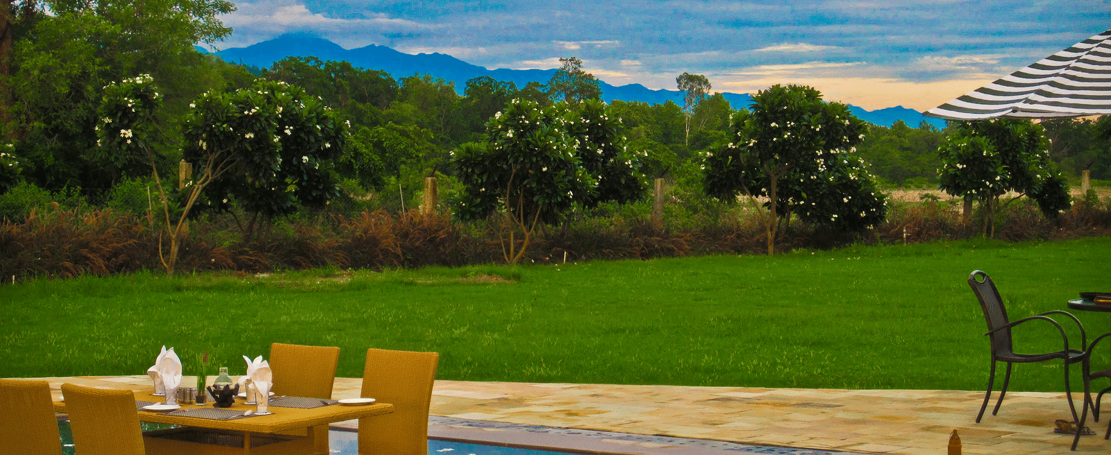 Full view shot of a lush green expansive lawn with a dining area half submerged in the swimming pool in the foreground - Ivory Destinations