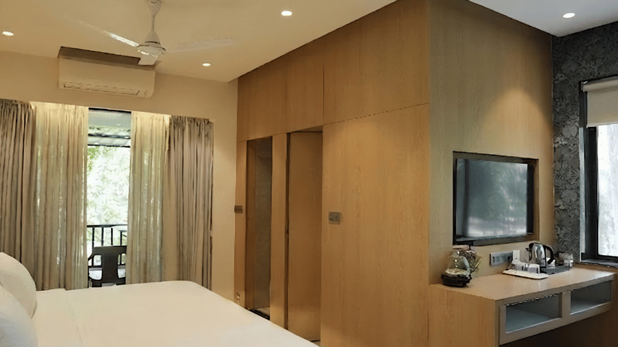 A bedroom shows a king size bed next to floor to ceiling wooden panelling, a vanity desk with a mirror, and a balcony view at Bougainvillea, Alibaug.
