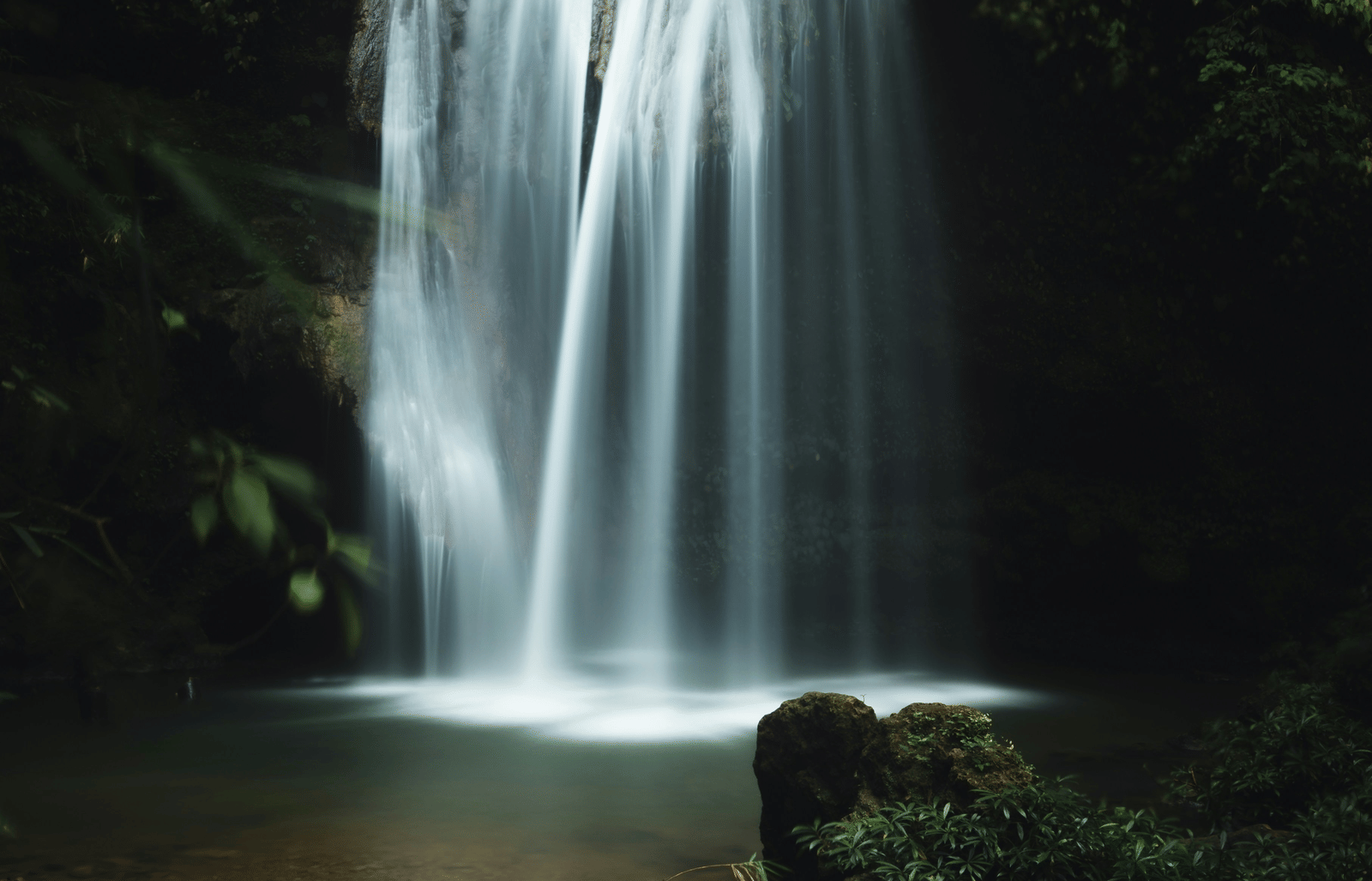 A long-exposure photograph of a multi-tiered waterfall cascading into a dark, foliage-rimmed pool.