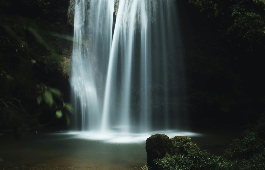 A long-exposure photograph of a multi-tiered waterfall cascading into a dark, foliage-rimmed pool.
