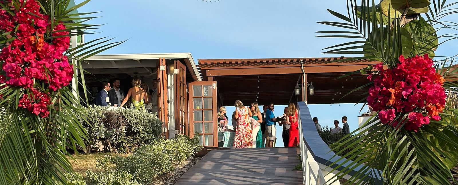 An wooden pathway leading to the cottage style courtyard where people have gathered to celebrate a wedding. The entrance of the decorated with branches, leaves, and flowers. 