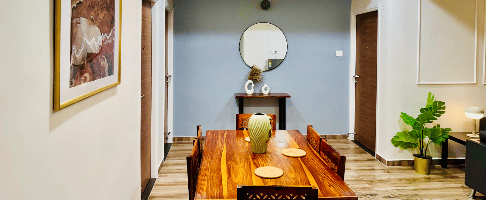 Dining area and common space in the Coral 4BHK at Kove Smart Stays featuring its wooden dining table and chairs placed at the centre of a common space that leads to doorways, and walls that are adorned with paintings and set-ups.