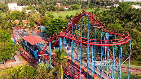 Aerial view of the Roller Coaster's red track on a blue steel frame, a red-roofed station, palm trees and the sea beyond at MGM Dizzee World, Chennai.