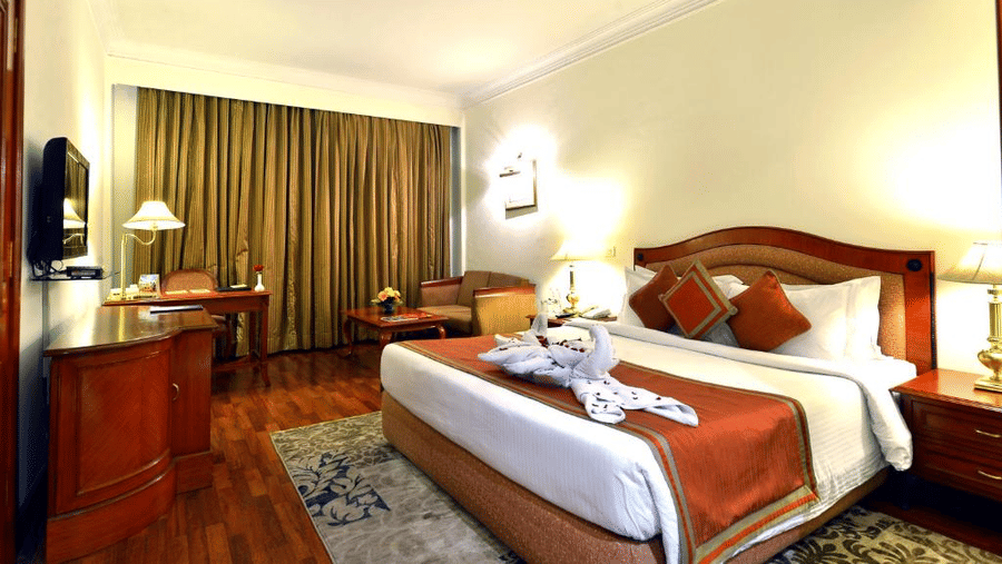 Standard Room at The Bristol Hotel, Gurgaon with a king-size bed, rust-coloured runner, a towel swan decoration, a wooden desk, an armchair, and curtains.