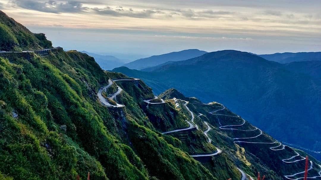 Scenic mountain road with hairpin turns winding through a sloping landscape, framed by majestic mountains and a vibrant sunset sky