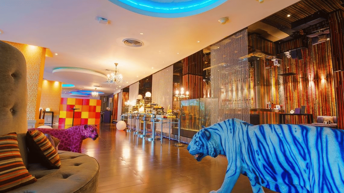 The interiors of Flava Rooftop bar, Bangkok, featuring warm ambient lighting, colourful wall art, plush seating, and a glowing blue tiger sculpture near the entrance | Night Hotel Bangkok