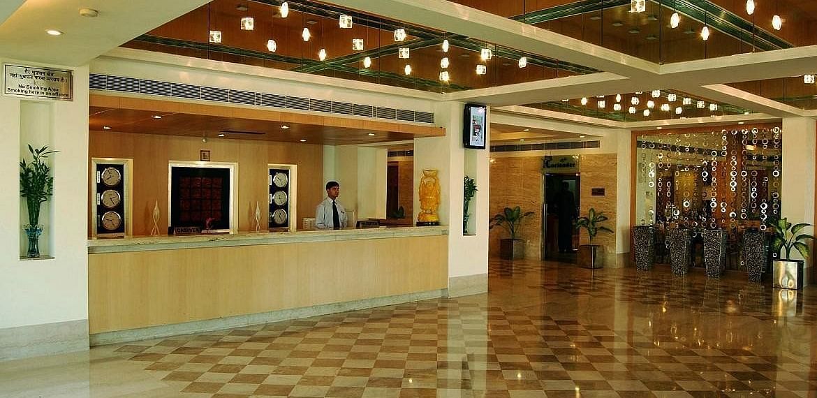 A well-lit hotel lobby with a check-in counter, receptionist, and a spacious polished marble floor with decorative lighting above - Lords Plaza Ankleshwar