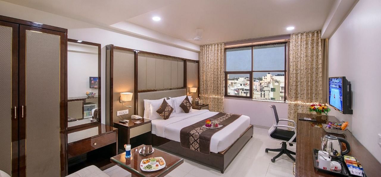 A hotel room at Lords Inn Porbandar, contains two single beds, a seating area, a television, and a view through the window.