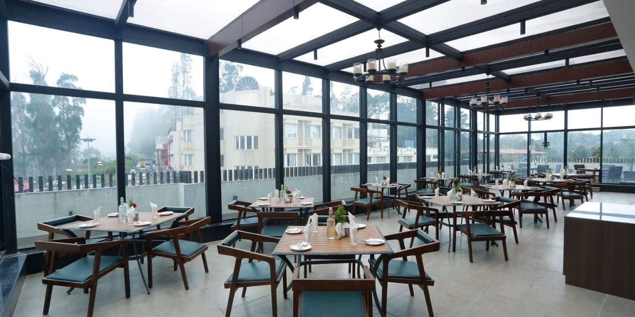 A rooftop dining area with glass walls and ceiling, offering stunning views and arranged tables and chairs for guests at The Hamuse Hotel, Kodaikanal.