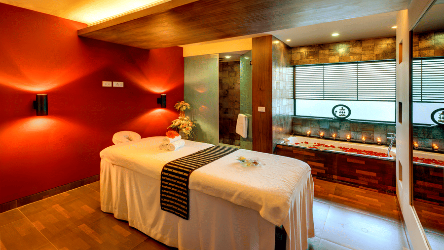 An overview of Sparoh Spa in Kolkata with a massage bed, a bathtub filled with water and rose petals, tea light candles next to the tub and aesthetic lighting at Kenilworth Hotel, Kolkata.