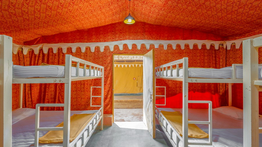 Wide-angle view of dormitory with multiple white bunk beds, orange decorative ceiling, brick walls, and bright multicoloured bedding arrangements.