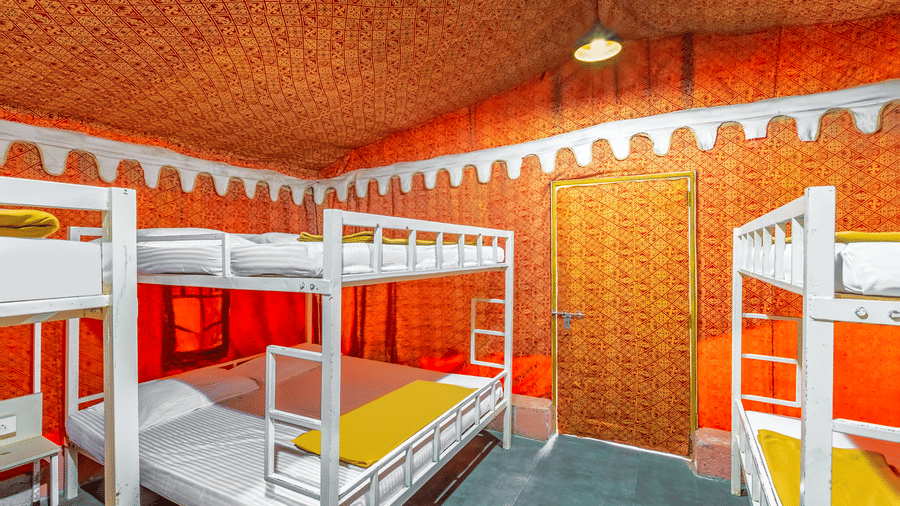 Side-angle dormitory view showing rows of white bunk beds under a vibrant orange ceiling with brick wall backdrop and colourful bedding.