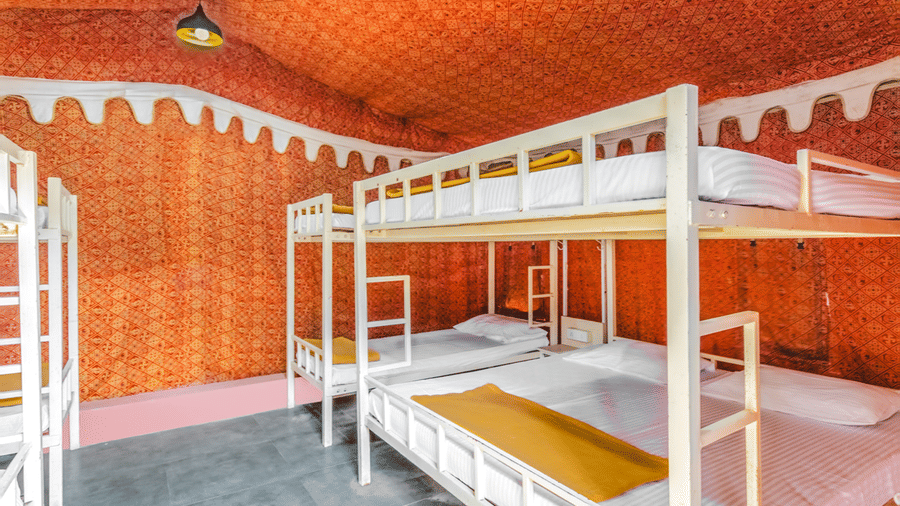 Close-up view of white metal bunk beds in dormitory with orange patterned tent ceiling, brick walls, and colourful mattresses on each bunk.