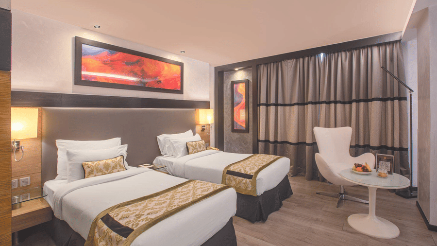 Modern hotel twin room featuring two single beds with gold runners and a small seating area.