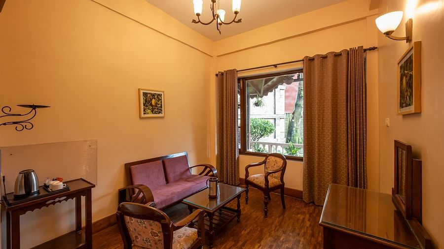 Cosy sitting area at The Pavilion, Nainital with purple sofa, wooden chairs, glass coffee table, and large window overlooking mountain views.