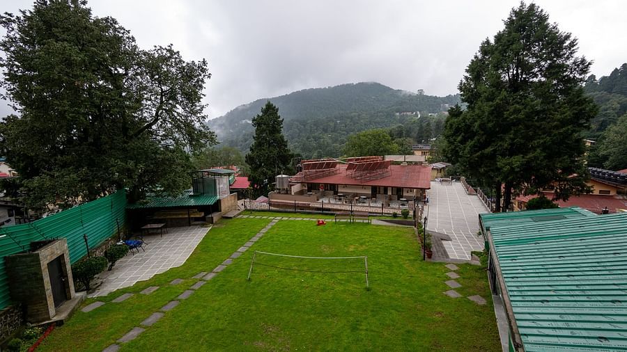 Expansive green lawn at The Pavilion, Nainital surrounded by forested hills, featuring volleyball net and stone pathways with mountain backdrop.