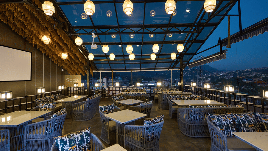An outdoor or rooftop restaurant with tables and chairs under a glass ceiling, illuminated by hanging glass globe lights, with a nighttime view of a city or landscape in the background | Voyage Eastin Suites